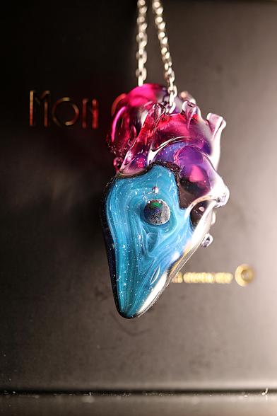 L’image montre un pendentif en verre en forme de cœur anatomique suspendu à une chaîne en argent 925. Le cœur est une pièce unique, réalisée en verre soufflé et sculpté au chalumeau.
Sa partie supérieure est composée de verre translucide aux teintes de rose et de violet, avec des détails évoquant les veines et artères d’un véritable cœur.
 La partie inférieure du cœur est principalement bleue, avec des tourbillons et nuances rappelant une galaxie lointaine.
 Au centre de cette zone bleutée se trouve une petite sphère en verre aux reflets noirs et argentés, évoquant une planète flottant dans l’univers.
La surface du verre est brillante et légèrement texturée par de petites bulles d’air emprisonnées. L’ensemble est mis en valeur par un fond sombre, où l’on aperçoit en arrière-plan des lettres dorées partiellement visibles. L’éclairage doux et chaud met en évidence la transparence et les couleurs vibrantes du verre, créant un effet de profondeur et de mystère.