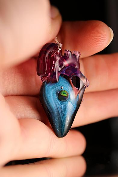 L’image montre un pendentif en verre en forme de cœur anatomique suspendu à une chaîne en argent 925. Le cœur est une pièce unique, réalisée en verre soufflé et sculpté au chalumeau.
Sa partie supérieure est composée de verre translucide aux teintes de rose et de violet, avec des détails évoquant les veines et artères d’un véritable cœur.
 La partie inférieure du cœur est principalement bleue, avec des tourbillons et nuances rappelant une galaxie lointaine.
 Au centre de cette zone bleutée se trouve une petite sphère en verre aux reflets noirs et argentés, évoquant une planète flottant dans l’univers.
La surface du verre est brillante et légèrement texturée par de petites bulles d’air emprisonnées. L’ensemble est mis en valeur par un fond sombre, où l’on aperçoit en arrière-plan des lettres dorées partiellement visibles. L’éclairage doux et chaud met en évidence la transparence et les couleurs vibrantes du verre, créant un effet de profondeur et de mystère.