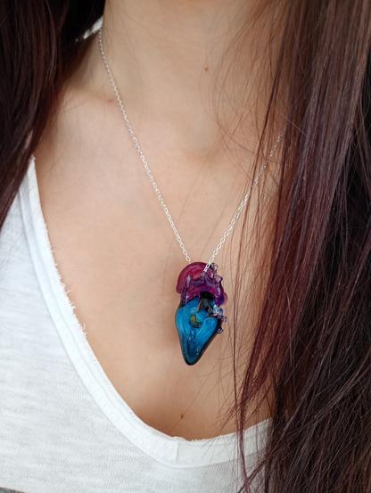 L’image montre un pendentif en verre en forme de cœur anatomique suspendu à une chaîne en argent 925. Le cœur est une pièce unique, réalisée en verre soufflé et sculpté au chalumeau.
Sa partie supérieure est composée de verre translucide aux teintes de rose et de violet, avec des détails évoquant les veines et artères d’un véritable cœur.
 La partie inférieure du cœur est principalement bleue, avec des tourbillons et nuances rappelant une galaxie lointaine.
 Au centre de cette zone bleutée se trouve une petite sphère en verre aux reflets noirs et argentés, évoquant une planète flottant dans l’univers.
La surface du verre est brillante et légèrement texturée par de petites bulles d’air emprisonnées. L’ensemble est mis en valeur par un fond sombre, où l’on aperçoit en arrière-plan des lettres dorées partiellement visibles. L’éclairage doux et chaud met en évidence la transparence et les couleurs vibrantes du verre, créant un effet de profondeur et de mystère.