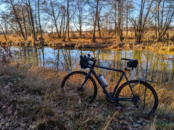 Rower szutrowy stojący wśród suchej trawy na samym brzegu niezbyt szerokiej rzeki. Na drugim brzegu drzewa, a za nimi łąki.