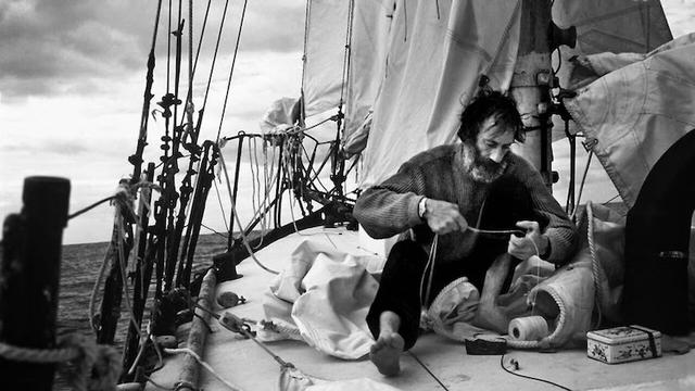 A man with unruly hair and a beard, Bernard Moitessier, sits on the deck of a sailboat at sea, sewing a sail. He doesn't wear expensive foul weather gear, just a jumper and loose pants,  barefoot.