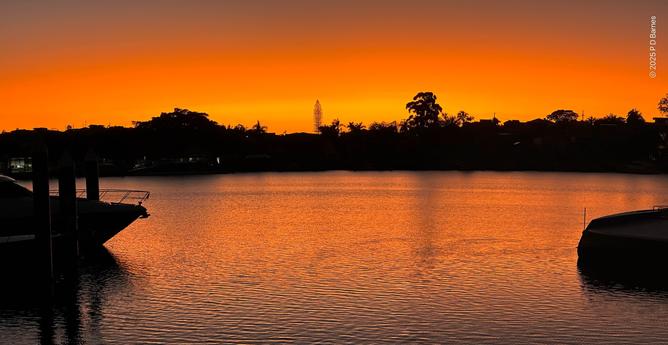 The Brisbane river at dawn. An intense orange glow lights the horizon, creating stark silhouettes of trees and buildings on the opposite bank. The river's surface is calm but rippled, blurring reflections but still lit up orange from  the sky. In the foreground the prow and stern in silhouette  of two boats moored on the near bank are at left and right respectively.