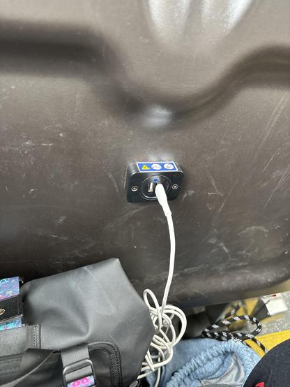 A white charging cord is plugged into a small rectangular plug receptacle on the back of a grey metal seat.