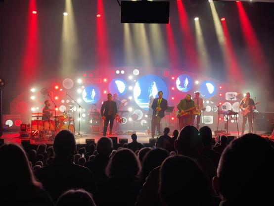 The band on stage, playing various instruments. The stage is illuminated with red and yellow spotlights and fog, creating a vibrant atmosphere. An audience is visible in the foreground, enjoying the show.