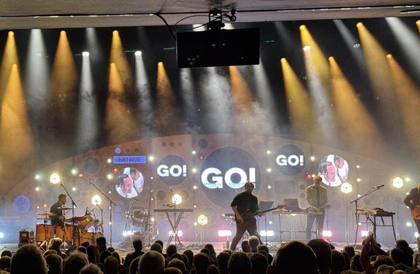The band in the middle of playing Go! There are yellow and white spotlights in the ceiling, shining down through smoke onto the stage