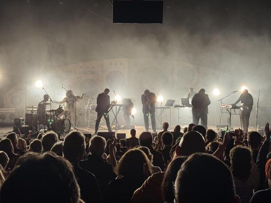 The four main members of the band, stationary at the end of a song, backlight by bright white lights shining through on-stage smoke
