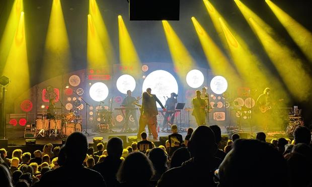 The band on stage playing various instruments, including drums and keyboards. Bright yellow stage lights shine down, illuminating the performer. Behind them is a large reproduction of an aircraft control panel with integrated screens and lights