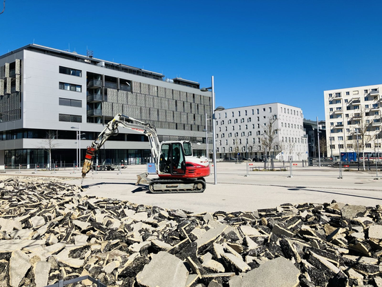Ein Bagger mit einem Presslufthammeraufsatz steht auf einem Platz aus Beton und entsiegelt. Im Vordergrund sieht man eine große Fläche bereits aufgerissenen Belags. Bei dem Platz handelt es sich um den Maria-Trapp Platz in Seestadt, im Hintergrund sieht man die Häuser an der Sonnenalle. Der Himmel ist strahlend blau und steht in einem starken Kontrast zur sonst grauen Architektur
