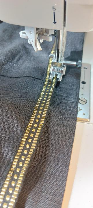 Un tissu en lin noir sous un pied de machine à coudre en train de fixer un galon noir et or