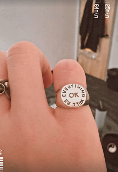 A silver ring on Ruibo's pinky finger. It has a flat, round top with the words 'Everything will be OK' written on it. 'OK' is at the center with the rest written in a circle around it.