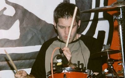 A man with short dark hair and wearing a grey t-shirt with long black sleeves, sitting behind a red and chrome drum kit, holding a pair of drumsticks in his hands, staring at his drums with a look of concentration on his face.
A microphone is mounted on the drum in front of him, with several cymbals on stands to his left, on the right of the frame.
Behind him is a red logo with a black and white ink drawing of a face in a helmet.