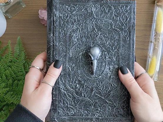 A5 notebook in silver color with a 3D clay cast of a crow skull. The notebook is black and made to look silver with decorative frame and corners.