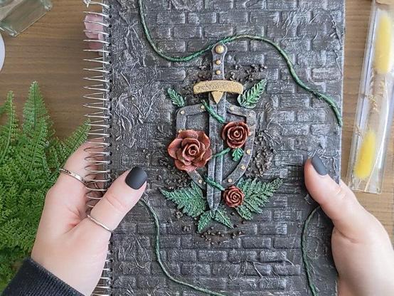 Cover of a spiral notebook with brick like relief background. There's a shield and a sword in silver and gold colors with three red acrylic roses glued on top. There are green air dry clay fern leaves framing the shield. Three strings of twine painted green are scattered around the perimeter. There is also a bit of decorative gravel in the nooks around the shield, painted slightly brown.