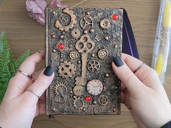 The cover of a A6 steampunk-inspired notebook in bronze color. There are embellishments on top, like a key, cog wheels, buttons and a clock. There are three ruby red gems scattered around the front.