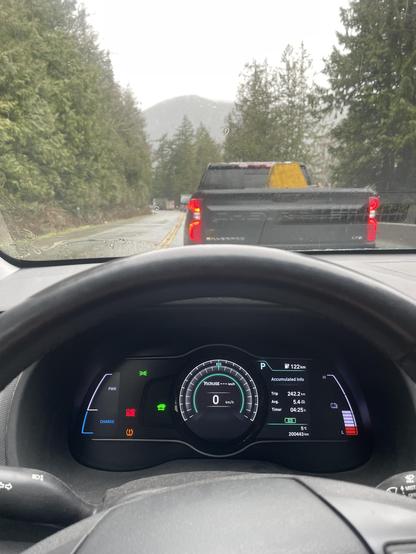 A picture of the front of the windshield looking at the dash of the car it is a digital display with various numbers for distance traveled, and the odometer which is at zero. The battery level is on the right and is just under half full out the windshield is ablack truck on the highway, which is surrounded by trees and mountain and Distance.