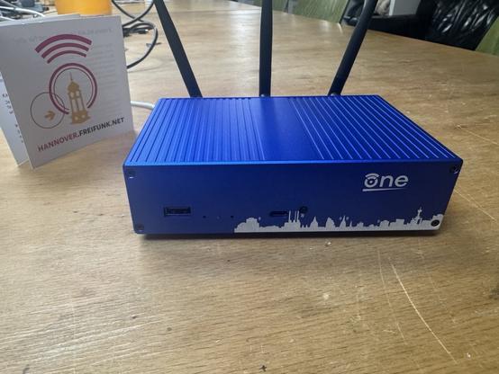 Ein OpenWrt One Router mit eloxiertem Gehäuse, in den das Logo "OpenWrt One", sowie die Freifunk Skyline Hannovers graviert wurde.