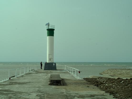 A green and white beacon on a pier by a lake, a person standing next to it, looking small in comparison.
