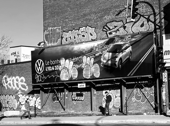 A person standing on a sidewalk in Montreal below a big publicity panel for a car brand but covered with tags. The photo is in black and white.