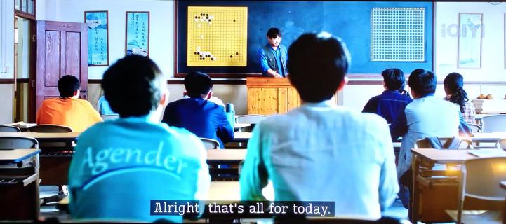 Paused TV screen of a Go classroom, all students facing away from the camera towards the teacher and the blackboard. The closest student on the left has a blue shirt on that says "Agender" underlined in big letters across the back.
