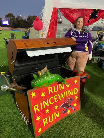 A woman in a Relay for Life with two stuffed dragons on her shoulders next to a giant luggage chest with teeth and lots of little legs. In front of the chest is a red and gold sign with the lettering Run Rincewind Run. There is an inflatable crocodile resting in the luggage's open mouth.
