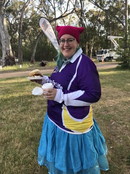 A woman wearing glases, a pink crochet beanie, a purple Relay for Life shirt, a blue skirt and a blue petticoat. She is holding a bacon and egg sandwich in one hand and a coffee in the other.