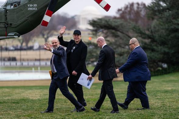Photograph of four men in black or dark blue suits walking in a line towards a helicopter on a grassy lawn. The men are all ethnically white and seemingly middle-aged. The first two men face the camera with smiles and waving hands. The second two look down and seemingly do not acknowledge the camera. A portion of the helicopter’s fuselage and tail rotor are visible in the upper left of the image’s frame. The photograph is credited to Eric Lee for The New York Times.
