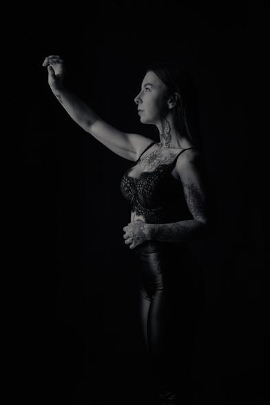 A black-and-white portrait of a woman with long hair, tattoos covering her arms, neck, and chest, wearing a lace bralette and high-waisted leather pants. She stands in profile against a dark background, raising one arm as if reaching for something unseen. The lighting casts dramatic shadows, emphasizing her tattoos and strong, elegant posture.