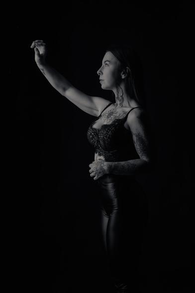 A black-and-white portrait of a woman with long hair, tattoos covering her arms, neck, and chest, wearing a lace bralette and high-waisted leather pants. She stands in profile against a dark background, raising one arm as if reaching for something unseen. The lighting casts dramatic shadows, emphasizing her tattoos and strong, elegant posture.