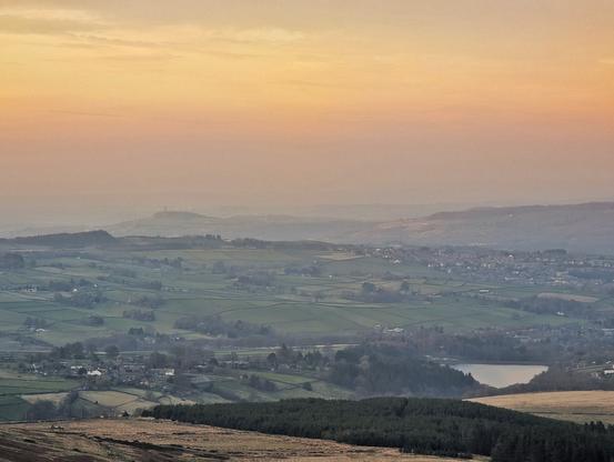 Moorland dropping away to rural landscape of fields, scattered housing, a reservoir, patches of wooded areas and copses, with more built-up areas beyond.  The view gets progressively mistier with distance.  A far salience looms with a tower atop