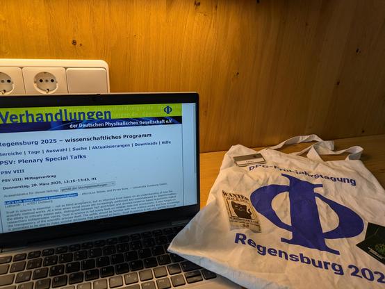 Aufgeklappter Laptop der #DPGR25-Programminformation für den Beitrag "Let's talk about science communication!" zeigt, Daneben ein hingelegter Tagungsbeutel mit Stickern und Namensschild