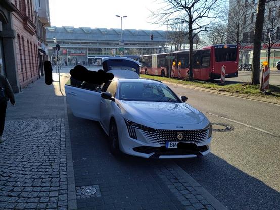 Ein weißer Pkw parkt halb auf dem Radweg, der durch einen Kantstein von der Straße getrenmt ist, und halb auf der Straße. Die hintere Tür ist geöffnet, sodass der Radweg vollständig versperrt ist. Die Heckklappe ist geöffnet, es werden Koffer eingeladen. Das Kennzeichen des Pkw ist unkenntlich, nur die ersten beiden Buchstaben "BW" sind zu lesen. Die Personen am Pkw sind auch unkenntlich gemacht.