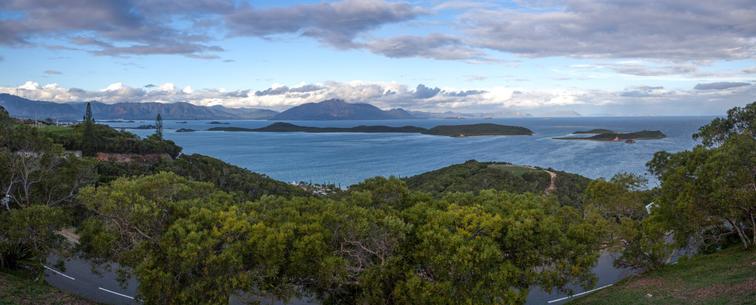 A panoramic view featuring a coastline with several small islands in the ocean, surrounded by lush greenery and distant mountains under a cloudy sky.
