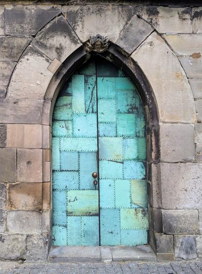 A close-up shot shows an arched doorway set into a weathered stone wall. The archway is constructed from large, roughly hewn grey stone blocks, showing signs of age and weathering. The keystone above the arch is adorned with a carved, slightly grotesque face.
The door itself is made of metal panels, likely copper or a similar alloy, now heavily patinated to a verdigris hue with shades of blue and green. The panels are rectangular and appear to be riveted together, creating a patchwork effect. A vertical seam runs down the centre where the two door leaves meet. A simple, round, brass-coloured doorknob is positioned on the right leaf. The bottom of the doorway meets a cobbled stone surface. The overall impression is of a historic and substantial entrance.
