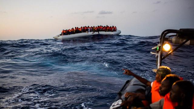 Ein großes, graues Gummiboot mit hunderten Menschen mit orangenen Rettungswesten auf dem Kamm einer Welle im Meer, im Vordergrund drei Schwarze Personen in einem Boot mit Scheinwerfer (das nur teils zu sehen ist), einer zeigt auf das Schlauchboot.