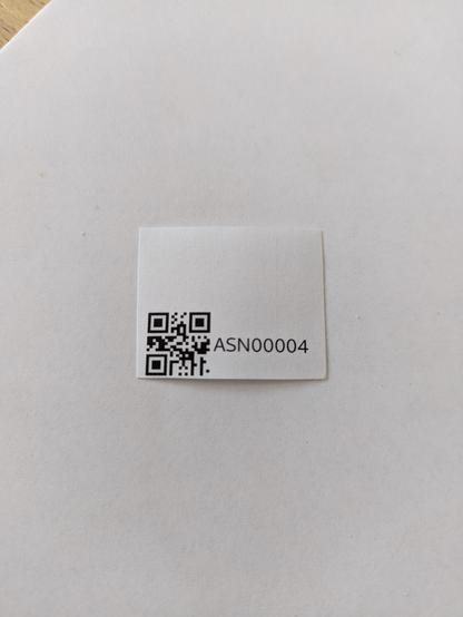 Ein kleines Label vor einem weißen Hintergrund. Das Label besteht aus einem QR-Code und einer Seriennummer, in diesem Fall ASN00004.