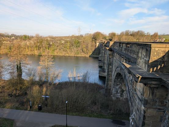 A view of the stone Aqueduct over the wide river. Either side of the river are grassy banks and lots of beautiful trees. Above is blue sky and wispy white clouds.