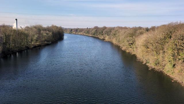 A view of the wide river from the Aqueduct with trees lining the sides of it. Above it blue sky and whispy clouds. On the right a factory tower of some sort can be seen.