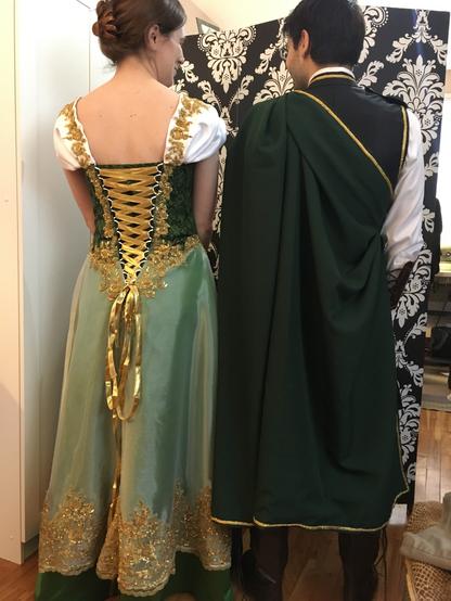 Back view of a woman and man.
Her hair is clipped up to show the open neckline of the dress. Bodice corset made in green cotton print with added sleeves in white satin.the skirt is a gold organza over green satin. Gold beaded lace trims the edges of the neckline, looped corset v back and the hem ove the skirt. Gold ribbon laces up the back opening.
He has a dark green semi cloak over a black vest, both trimmed in gold braid.