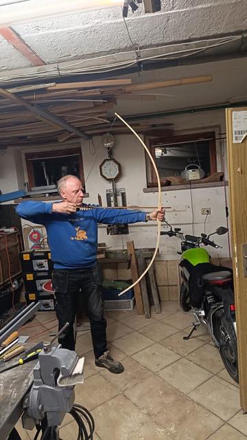 Der lehrling zieht den halbfertigen Bogen mit einem eingelegten Pfeil aus und wird von der Seite aus fotografiert. Das auch zur Kontrolle, wie sich der Bogen verformt und biegt