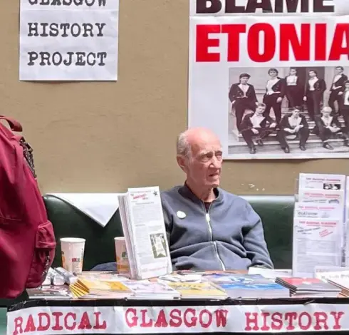 John sitzend hinter einem Infotisch.