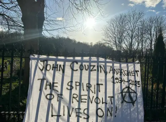 An einem Zaun hängt ein Transparent mit dem Text "John Couzin, Anarchist, 1934-2025 - The Spirit of Revolution lives on!"