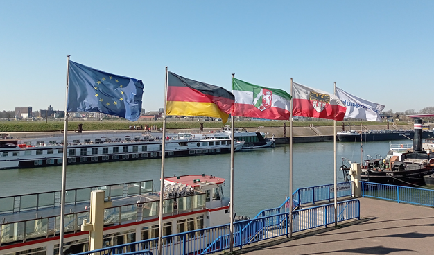 5 Flaggen an der Schifferbörse wehen in Wind und Sonnenschein. Von links nach rechts Europa, Deutschland, NRW, Duisburg und Duisport.

Im Hintergrund die Gerhard Mercator, Hafenrundfahrtsschiff der weißen Flotte, rechts ein Teil der Oscar Huber, Museumsschiff. Auf der anderen des Kanals 2 Ausflugsschiffe und weiter rechts ein Tanker angelegt an der Mercatorinsel.