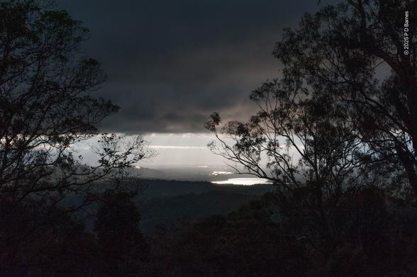 High view down into a valley and plains. Most of the sky is obscured by a very heavy, dark bank of cloud, creating an almost night-time dimness from the foreground to a long way out on the plain. The cloud has a very distinct edge, and beyond that there is bright sunshine, which reflects off a dam, and the very hazy plains beyond, creating a thin band of bright light between cloud and ground. The dominant feature in the image however is the silhouettes of two large, relatively sparse trees, whose filigree-like outlines occupy most of the left- and right-hand sides of the image.