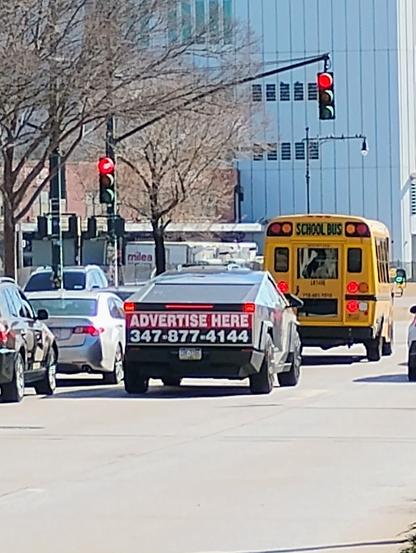 A CyberTruck behind a school bus with white texts, namely "ADVERTISE HERE" on red background and a phone number on black background, on the back trunk.