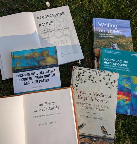 The open books on the meadow. The titles are: "Reconceiving Nature: Ecofeminism in Late Victorian Women's Poetry" by Patricia Murphy; "Post-Romantic Aesthetics in Contemporary British and Irish Poetry" by Stefanie John; "Can Poetry Save the Earth?" by John Felstiner; "Birds in Medieval English Poetry" by Michael J. Warren; "Poetry and the Anthropocene" by Sam Solnik; "Writing on sheep" by William Welstead