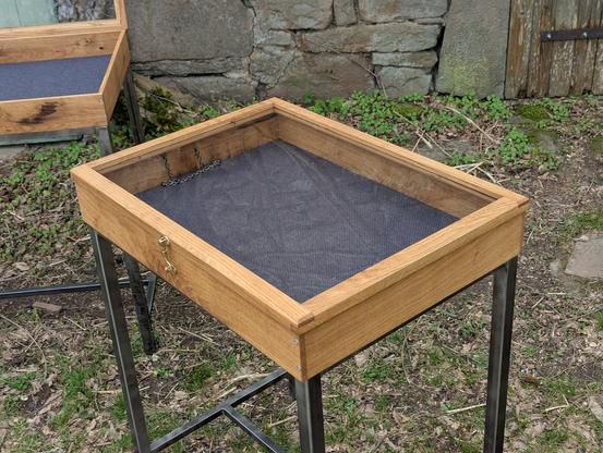 Une vitrine d'exposition avec un cadre en bois et une vitre transparente sur le dessus. Elle est montée sur des pieds en métal noir. La vitrine est fermée, révélant un intérieur vide avec un fond gris anthracite et une chaînette noire. À l'arrière-plan, on peut voir une deuxième vitrine.