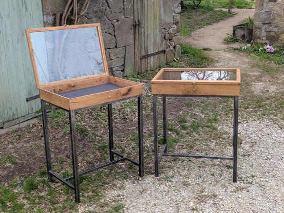 Deux vitrines d'exposition. Chaque vitrine est composée d'un cadre en bois avec une vitre transparente sur le dessus. Elles sont montées sur des pieds en métal noir. L'une des vitrines est ouverte, révélant un intérieur vide avec un fond noir. Les vitrines sont placées sur un sol de gravier à l'extérieur, près d'un mur en pierre et d'une porte en bois verte.