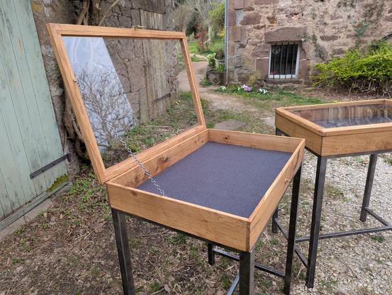 Une vitrine d'exposition avec un cadre en bois et une vitre transparente sur le dessus. Elle est montée sur des pieds en métal noir. La vitrine est ouverte, révélant un intérieur vide avec un fond gris anthracite. La vitrines est positionnée sur un sol de gravier près d'un mur en pierre et d'une porte en bois. À l'arrière-plan, on peut voir un chemin menant à une autre structure en pierre avec une petite fenêtre et une deuxième vitrine.