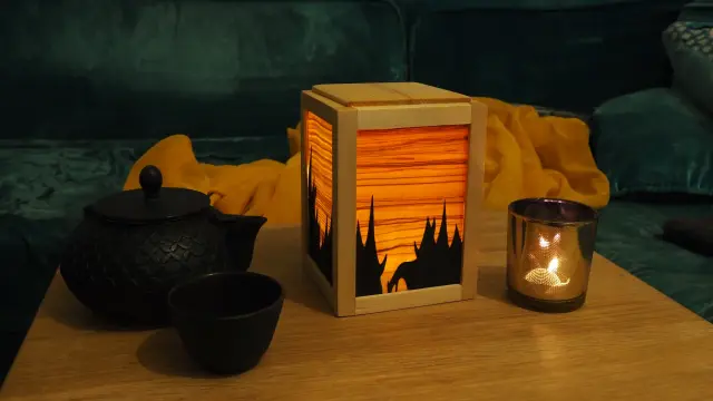 Photo d'une veilleuse en bois. C'est une sorte de cube. Chaque côté de la veilleuse est fait d'un cadre dans lequel se trouve de la marqueterie. La lumière passe à travers la fine feuille de bois et met en valeur le veinage du bois. Par endroit une deuxième couche de placage fait se dessiner une forêt de sapin en négatif.