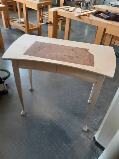 Photo d'un bureau dans un atelier. Il n'y presque aucune ligne droite : le plateau, les pieds et les tiroirs sont arrondis. Le plateau est marqueté avec une essence de bois foncé qui esquisse un sous main de bureau.